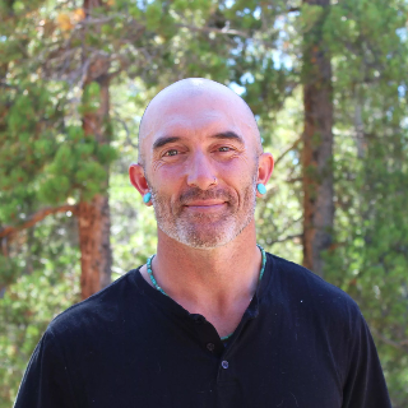 The image shows a middle-aged man with a shaved head and a neatly trimmed grey beard. He has a nose ring and turquoise earrings. He is wearing a black button-down shirt and a turquoise necklace. The background features a blurred forest setting with green trees. The man is looking directly at the camera with a slight smile.